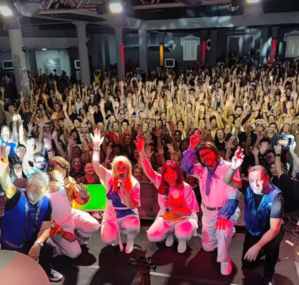 Banda acenando no palco com o salão lotado atrás, no show esgotado em Recife